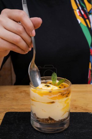 Téléchargez les photos : Manger un dessert. Vue rapprochée de la main d'une femme tenant une cuillère, ayant un dessert de mousse de fruits de la passion, avec de la farine d'avoine et des noix, servi dans un verre. - en image libre de droit