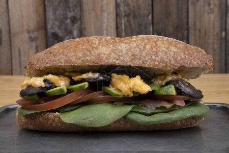 Foto de Un delicioso sándwich hecho con pan baguette, aguacate, champiñones, tomate y espinacas, en un plato de metal con fondo de madera. - Imagen libre de derechos