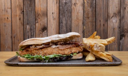 Foto de Sandwich gourmet. Vista de cerca de un sándwich multicapa con filete frito de pollo, tocino, queso y lechuga, con papas fritas. - Imagen libre de derechos