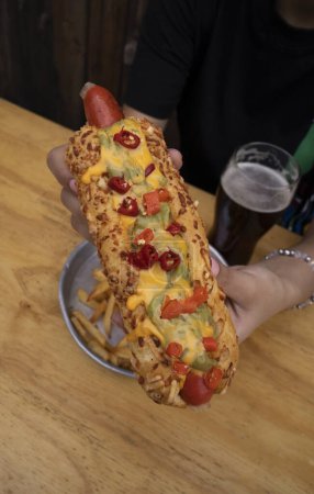 Téléchargez les photos : De la nourriture américaine. Vue rapprochée d'une femme tenant un hot-dog gastronomique avec guacamole, chilis et fromage cheddar. Des frites et une bière sur la table. - en image libre de droit