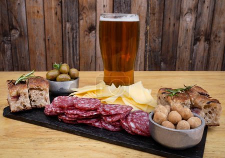 Photo for Picada. Closeup view of a dish with sliced salami, cheese, focaccia peanuts, green olives and a pint of beer on the wooden table. - Royalty Free Image