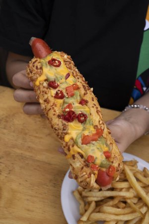 Foto de Comida rápida gourmet. Vista de cerca de una mujer comiendo un hot dog mexicano con guacamole, queso cheddar y jalapeños, con papas fritas y un vaso de cerveza. - Imagen libre de derechos