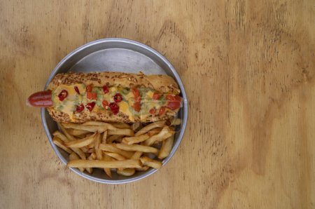 Foto de Comida rápida gourmet. Vista superior de un hot dog con guacamole, chiles, queso cheddar y papas fritas, en un plato de metal con fondo de madera. - Imagen libre de derechos