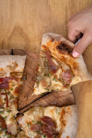 Photo for Eating pizza. Closeup view of a woman's hand serving a slice of pizza with tomato sauce, mozzarella cheese, crispy bacon and onions, on the wooden table. - Royalty Free Image