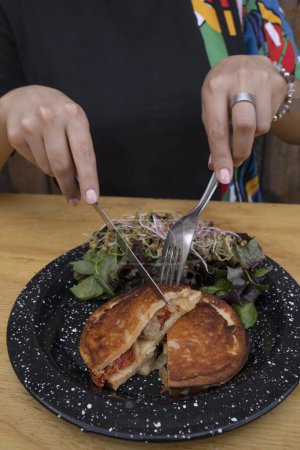Téléchargez les photos : Manger de la provoleta. Vue rapprochée d'une femme tenant un couteau et une fourchette, mangeant un fromage provolone grillé farci avec une salade verte fraîche. - en image libre de droit