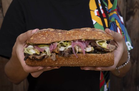 Foto de Mujer sosteniendo un delicioso sándwich hecho con pan de baguette, lechuga, encurtidos de cebolla roja y carne a la parrilla. - Imagen libre de derechos
