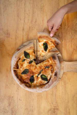 Photo for Traditional Margherita pizza, Top view of a female hand serving a slice of pizza with mozzarella and parmesan cheese, tomato sauce and basil, in the restaurant. - Royalty Free Image