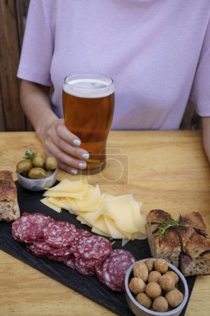 Photo for Picada. Closeup view of a woman drinking a beer and having cold cuts, such as sliced salami, cheese, focaccia peanuts and green olives. - Royalty Free Image