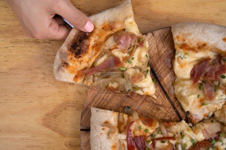 Foto de Comiendo pizza. Vista de cerca de la mano de una mujer que sirve una rebanada de pizza con salsa de tomate, queso mozzarella, tocino crujiente y cebolla, en la mesa de madera. - Imagen libre de derechos