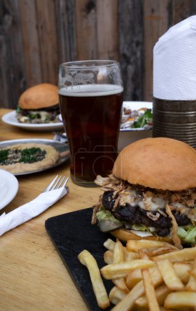 Photo for Selective focus on a hamburger with fries. Other dishes with burgers, hummus and a pint of beer in the background. - Royalty Free Image