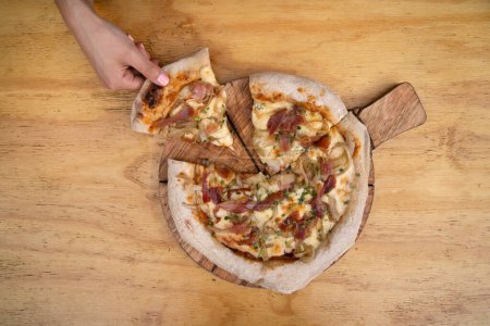 Foto de Vista superior de una mano sosteniendo una pizza hecha con salsa de tomate, queso mozzarella, tocino crujiente y cebolla, con un fondo de madera. - Imagen libre de derechos