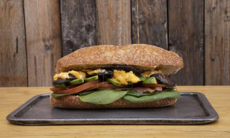 Foto de Un delicioso sándwich hecho con pan baguette, aguacate, champiñones, tomate y espinacas, en un plato de metal con fondo de madera. - Imagen libre de derechos