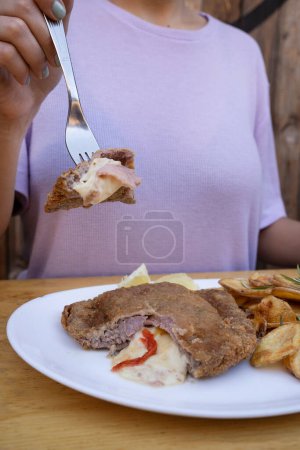 Foto de Mujer sosteniendo un tenedor, tomando una milanesa, bistec frito empanado, relleno con queso mozzarella, jamón y pimiento morrón con patatas fritas y limón, en un plato blanco. - Imagen libre de derechos