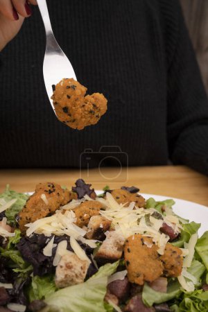 Photo for Closeup view of a woman holding a fork with a fried shrimp, eating a Caesar salad with lettuce, parmesan cheese, bread croutons and crispy bacon. - Royalty Free Image