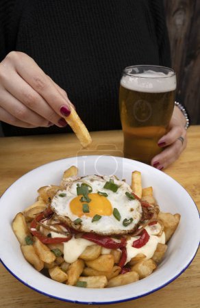 Téléchargez les photos : Une femme prenant une bière avec des pommes de terre frites avec un oeuf grillé, poivron rouge rôti, oignons et fromage à la crème dans un bol blanc. - en image libre de droit