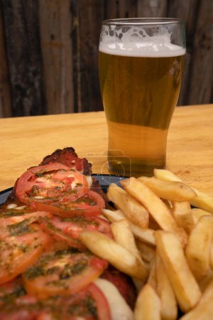 Photo for Milanesa napolitana. Closeup view of fried breaded steak with mozzarella cheese, tomato, garlic, basil, french fries and a glass of beer. - Royalty Free Image