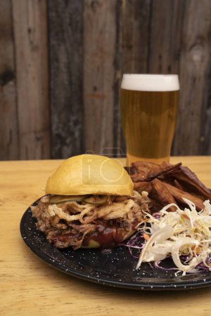 Photo for Closeup view of a pulled pork burger with bbq sauce, pickles and crispy onion. Fried sweet potatoes, coleslaw salad and a glass of beer, complete the dish. - Royalty Free Image