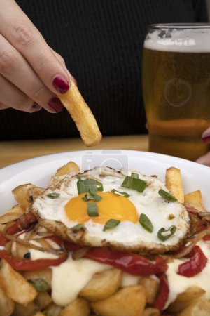 Photo for A woman having a beer with fried potatoes with a grilled egg, roasted red bell pepper, onions and cream cheese in a white bowl. - Royalty Free Image