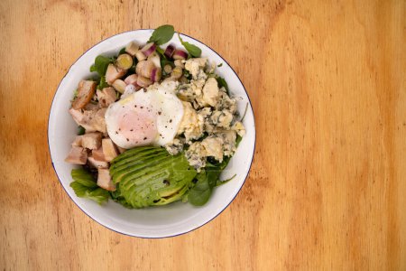 Photo for Top view of a fresh salad with poached egg, avocado, blue cheese, greens and grilled chicken breast, on the wooden table. - Royalty Free Image
