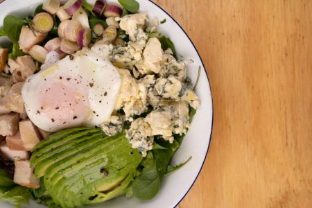 Photo for Top view of a fresh salad with poached egg, avocado, blue cheese, greens and grilled chicken breast, on the wooden table. - Royalty Free Image