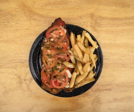 Photo for Milanesa napolitana. Top view of fried breaded steak with mozzarella cheese, tomato, garlic, basil and french fries, in a black dosh on the wooden table. - Royalty Free Image