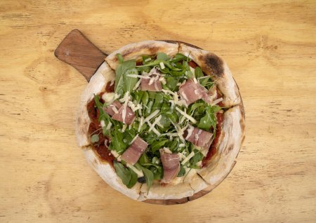 Foto de Vista superior de una pizza con rúcula, jamón curado, queso parmesano, queso mozzarella y salsa de tomate, sobre la mesa de madera. - Imagen libre de derechos