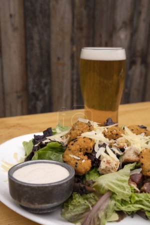 Photo for Closeup view of a Caesar salad with fried breaded shrimps, lettuce, parmesan cheese, bread croutons, crispy bacon and a glass of beer. - Royalty Free Image
