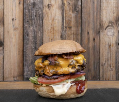 Monster-Burger. Nahaufnahme eines mehrschichtigen Burgers mit zwei Fleischmedaillons, Cheddar-Käse, Speck, Salat, Tomaten, Ketchup und Mayonnaise, auf dem Tisch mit Holzgrund.