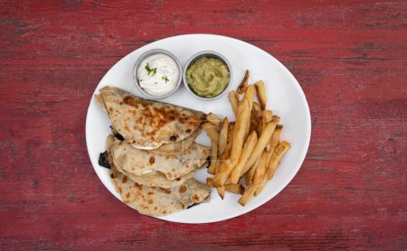 Photo for Mexican cuisine. Top view of quesadillas filled with cheese, french fries and dipping sauces, in a white dish on the restaurant red wooden table. - Royalty Free Image