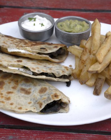 Photo for Mexican cuisine. Closeup view of quesadillas filled with cheese, french fries and dipping sauces, in a white dish on the restaurant red wooden table. - Royalty Free Image