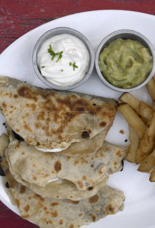 Photo for Mexican cuisine. Top view of quesadillas filled with cheese, french fries and dipping sauces, in a white dish on the restaurant red wooden table. - Royalty Free Image