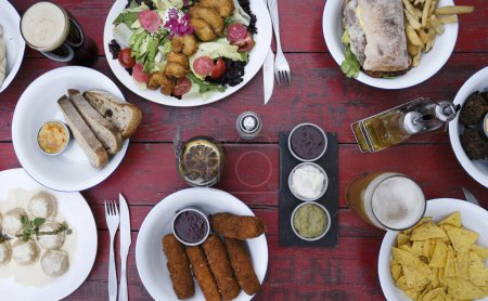 Photo for Banquet. Top view of different dishes on the restaurant wooden table. Food such as beer, salad, mozzarella sticks, nachos, shrimps, pasta and a sandwich with fries. - Royalty Free Image