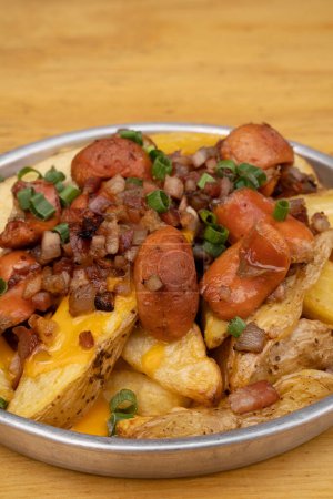 French fries. Top view of fried potatoes with sliced green onion, smoked sausages, crispy bacon and cheddar cheese sauce, in a metal dish on the wooden table.