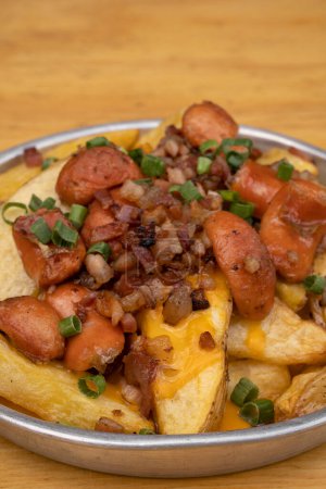 French fries. Top view of fried potatoes with sliced green onion, smoked sausages, crispy bacon and cheddar cheese sauce, in a metal dish on the wooden table.