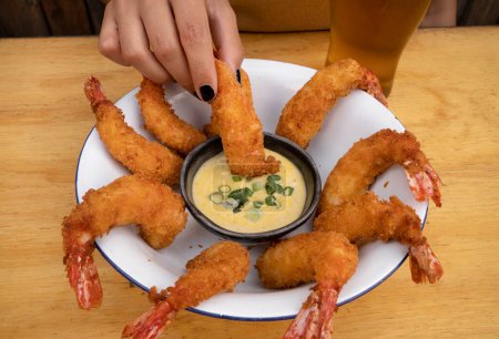 Closeup view of a woman's hand dipping a fried shrimp tail in a creamy sauce. 
