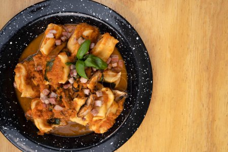Des pâtes. Vue du dessus de l'agnolotte farcie traditionnelle avec sauce tomate aux champignons, bacon et basilic dans un plat noir sur la table en bois.