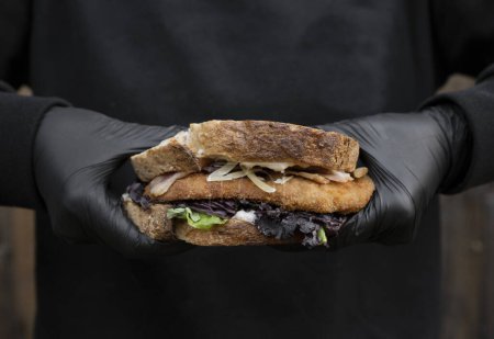 Chef hands holding a sandwich made with bread, parmesan cheese, lettuce, bacon and fried chicken steak, with a black background.
