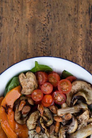 Top view of a nutritious salad with grilled vegetables. Vegetarian salad with grilled pumpkin, mushrooms, spinach, barley, almonds and cherry tomatoes, in a white bowl on the restaurant wooden table.