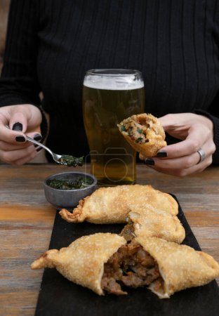 Photo for Eating empanadas at the restaurant. Closeup view of a caucasian woman having fried meat empanadas with traditional chimichurri sauce and a pint of beer - Royalty Free Image
