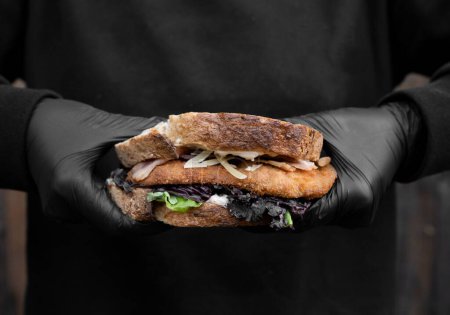 Küchenchef hält ein Sandwich mit Brot, Parmesan, Salat, Speck und gebratenem Hühnersteak mit schwarzem Hintergrund in der Hand.