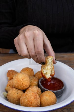 Eating cheese balls at the restaurant. Closeup view of a caucasian woman's hand, dipping a potato and mozzarella croquette in sweet chili dipping sauce