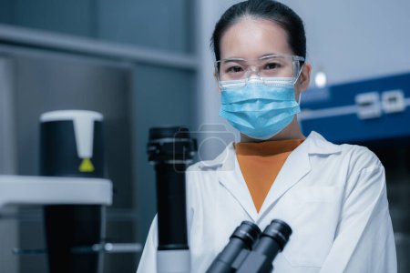 Photo for Portrait of a female researcher, a scientist wearing safety goggles and a mask working in the laboratory with a microscope to look at culture cells for bioassay. Research, medicine, biotechnology development. - Royalty Free Image