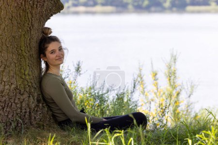 Téléchargez les photos : Une jeune femme brune assise dans une forêt verdoyante profite du silence et de la beauté de la nature qui veille sur un lac de forêt bleue lors d'une journée d'été. Photo de haute qualité - en image libre de droit