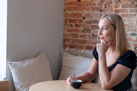 Téléchargez les photos : Une photo captivante d'une jeune femme blonde immergée dans ses pensées, assise dans un charmant café vintage moderne au cœur de la ville. Elle tient une tasse de café dans une main, tandis que le - en image libre de droit