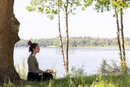Photo for A serene and tranquil moment captured in nature, featuring a young brunette woman sitting gracefully in the lush green grass next to a majestic tree. She faces the peaceful forest lake, her eyes - Royalty Free Image