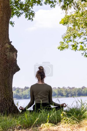 Photo for A serene and tranquil moment captured in nature, featuring a young brunette woman sitting gracefully in the lush green grass next to a majestic tree. She faces the peaceful forest lake, her eyes - Royalty Free Image