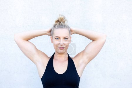 Capture the joy of relaxation and stretching with this footage. A young blonde woman stands against a serene concrete background, arms and hands gently behind her head and neck. With a content smile