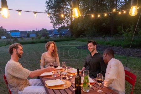 Eine multiethnische Gruppe tausender Freunde versammelte sich an einem Grill-Esstisch vor einem schönen Haus mit Lichtern in der Abenddämmerung. Junge Leute haben Spaß und essen Essen. Gartenfest-Feier in einem