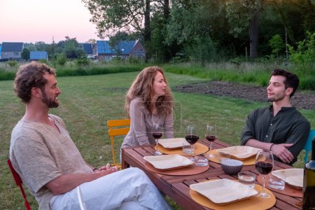 Photo for Three close European friends gather around a table in a serene garden as the sun sets. With glasses of wine in hand, they engage in a warm and relaxed conversation, savoring the beauty of the evening - Royalty Free Image