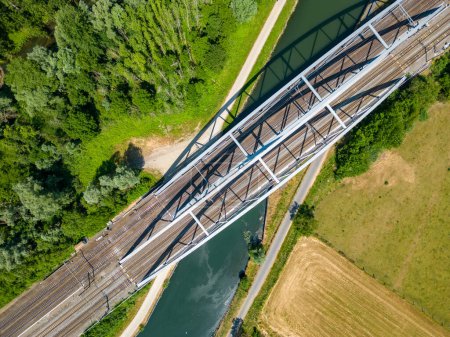 Photo for An awe-inspiring aerial perspective of a steel railroad bridge gracefully spanning over a serene river or canal. The intricate design and engineering marvel of this bridge provide a striking contrast - Royalty Free Image
